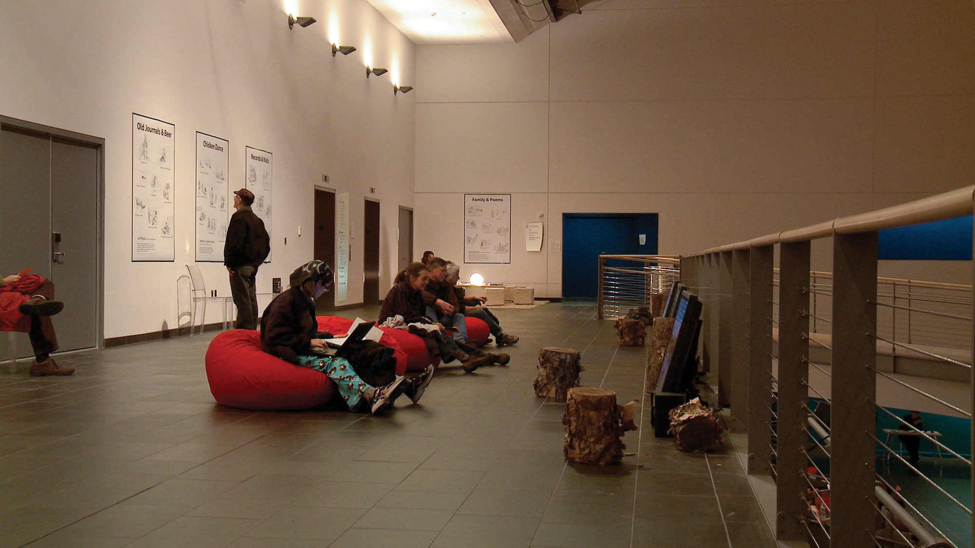 A group of about five people seated in red bean bag chairs on the floor of a hallway as other people mill about looking at posters hung on the walls. 