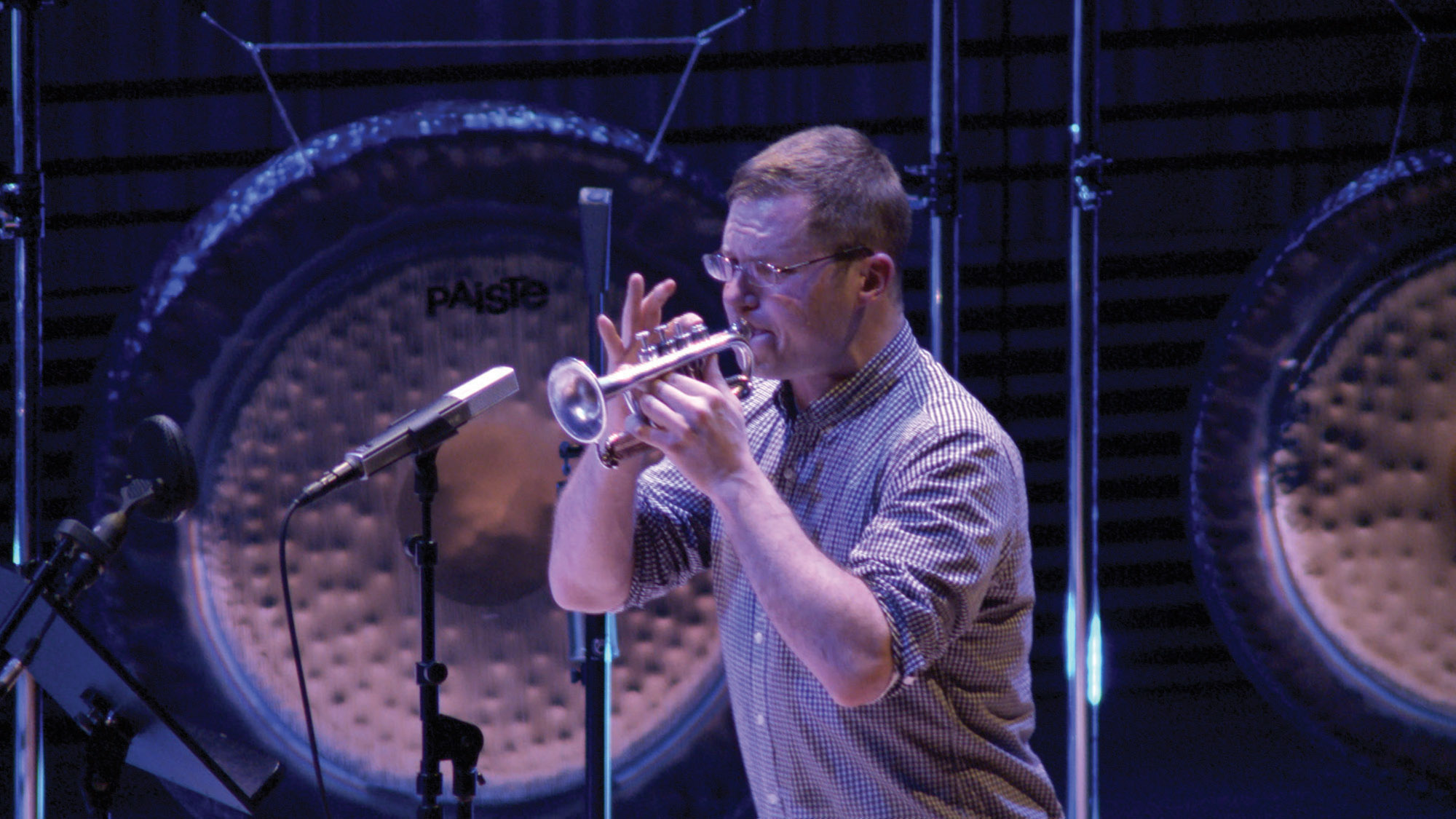 A white male playing a soprano trumpet into a microphone in front of a gong. 
