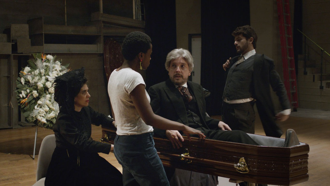 Two people wearing victorian style mourning clothes mill about a funeral scene as a man with gray hair dressed similarly sits up in a coffin talking to a Black female director wearing contemporary clothing. 