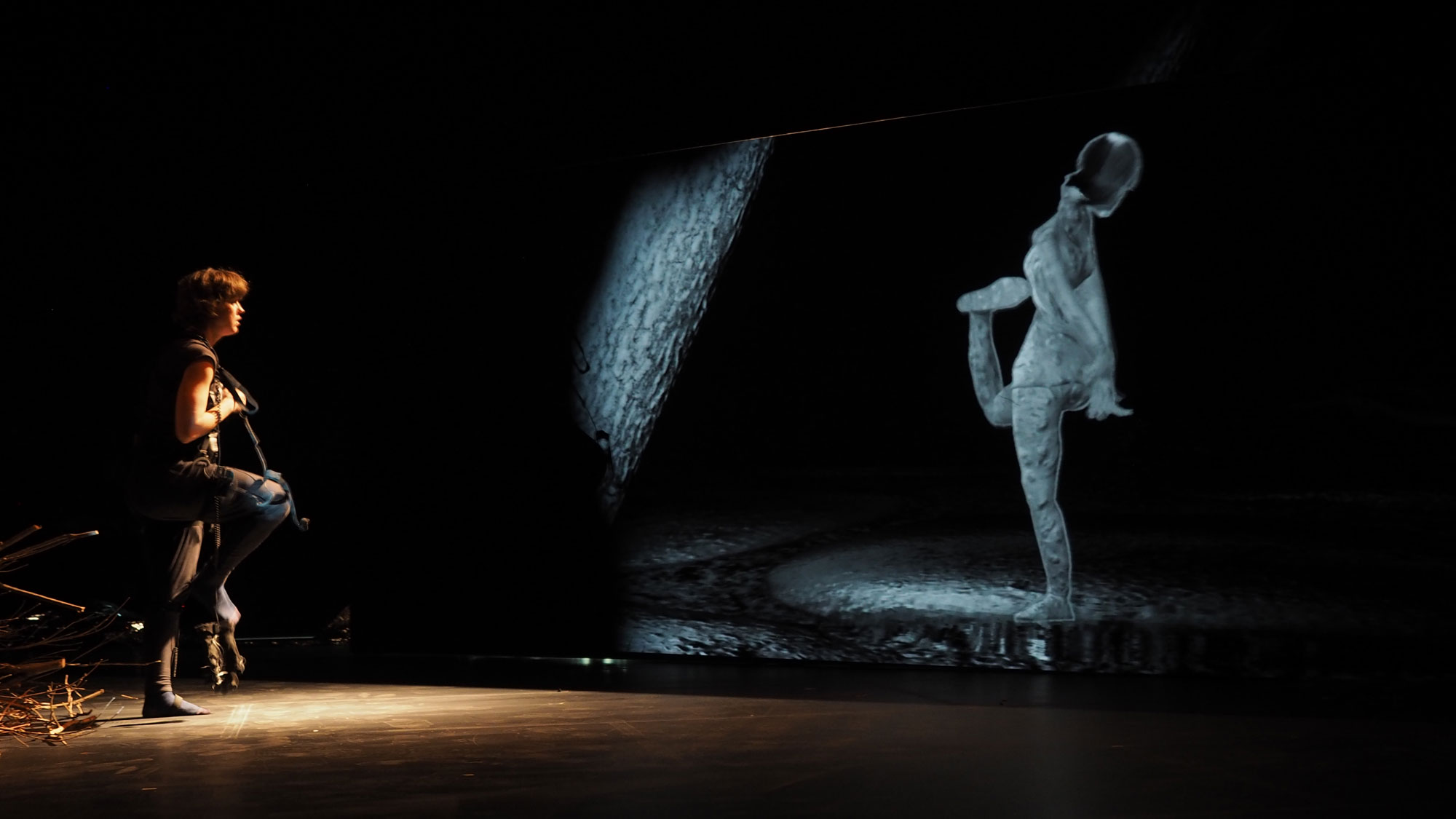 A woman wearing rigging stands on one leg as her image is projected on to a screen in front of her in a black box studio. 