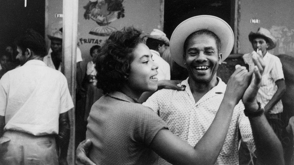 A Black Cuban couple wearing 1950's clothing dancing joyfully in a crowded street. 