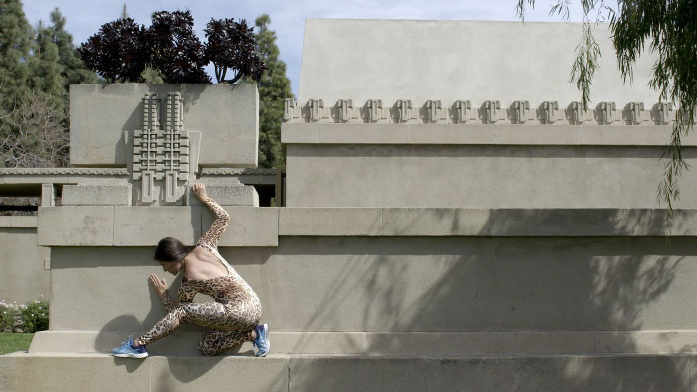 A woman wearing a cheetah print unitard and blue athletic sneakers crouching on a concrete wall. 