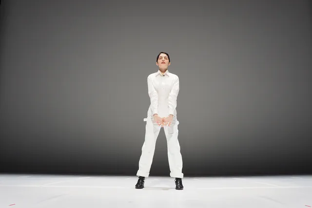 A woman wearing a white jumpsuit standing in front of a grey backdrop. 