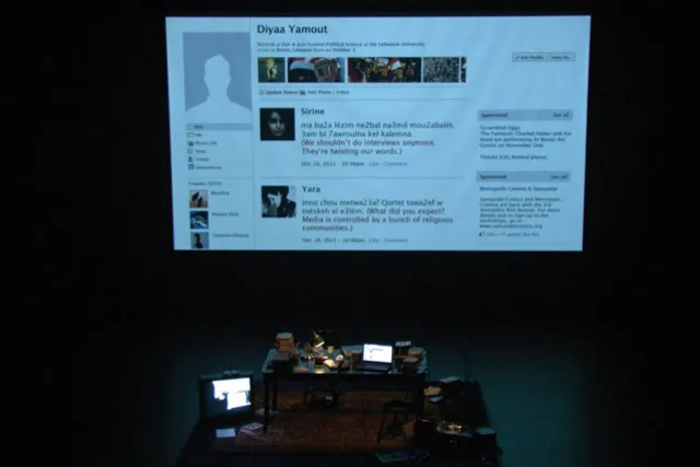 A black table cluttered with various office items in front of a projection of a facebook profile