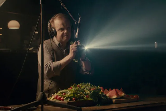 A man standing in front of a large salad and cut up watermelon, crinkling a piece of lettuce into a microphone. 