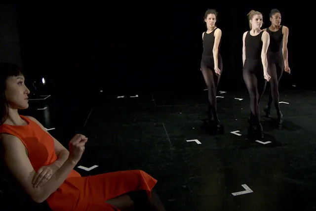 A woman in orange sits int he foreground with legs crossed as three other woman in black leotards walking in a V formation. 