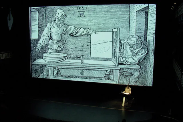 a woman giving a talk on a black stage in front of a projection of a 1500's sketch. 