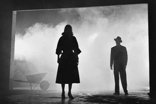 A man and woman wearing 1940's era clothing looking out into dense, gray fog with backs to the viewer.  