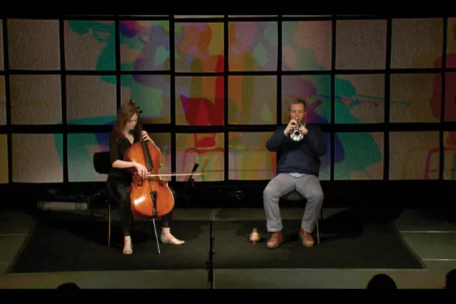 A woman playing the cello seated next to a man playing the trumpet in front of a grid of acoustic tiles with a rainbow shadow. 