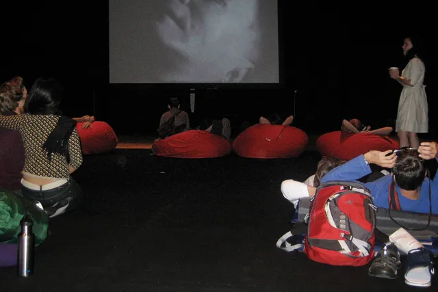 Various people seated in red bean bag chairs in a theater. A couple sits in the foreground in an embrace, a woman in a white dress finds her seat while holding a cup. 
