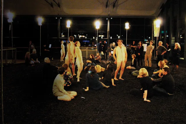 A small audience seated around four performers wearing white rompers standing thought out the space. 
