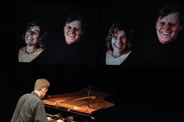 A man seated at a piano with back to the viewer as four projections of smiling people floor above him. 