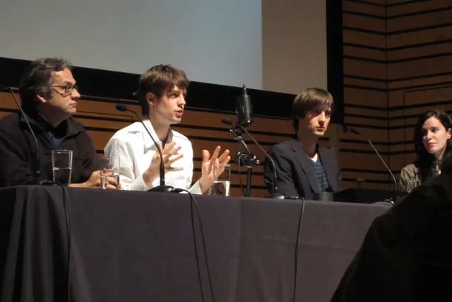 A panel of three men and one woman seated behind a table with a black table cloth. A man in a white shirt is speaking into a microphone with his hands outstretched. 