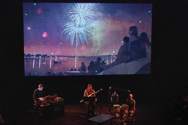 A group of three musicians playing on a dark stage lit by a projection of fireworks on a screen behind them. 
