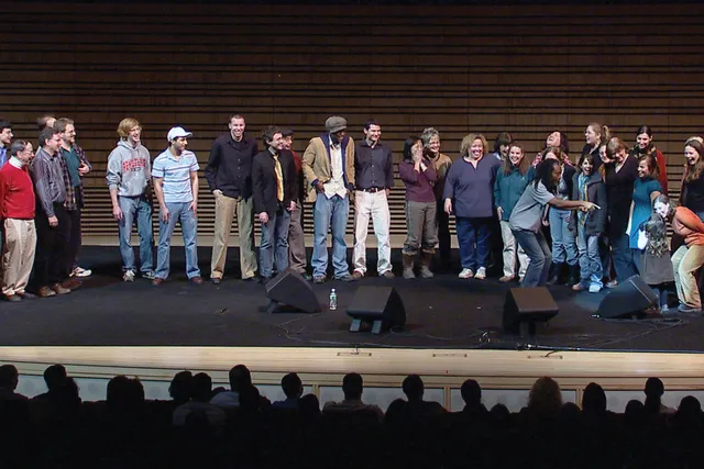 An assorted group of about 30 people standing on the concert hall stage in front of an audience. 