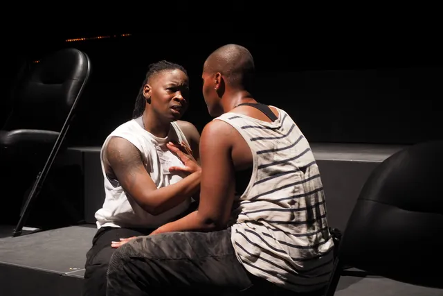 Two Black woman sitting facing each other on the steps of a black box theater. 