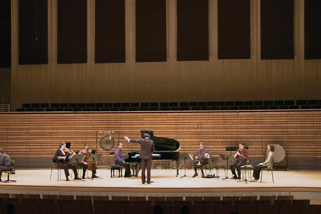 A small orchestra spread out across the concert hall stage as two men play chess in the corner of the stage. 