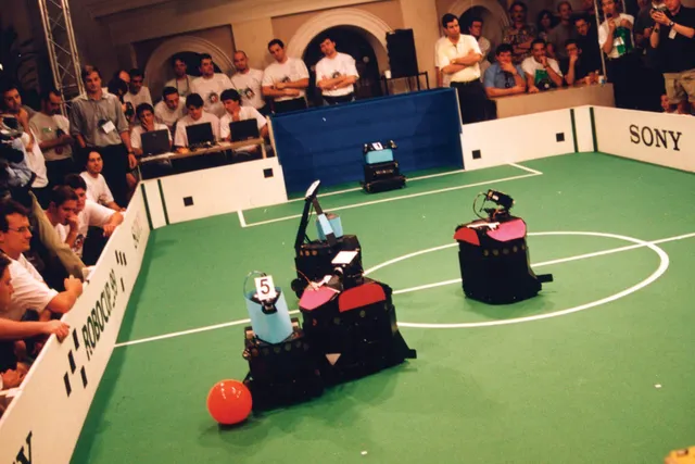 Six robots wearing blue or red on a pitch playing a form of soccer with a red ball surrounded by men controlling them. 