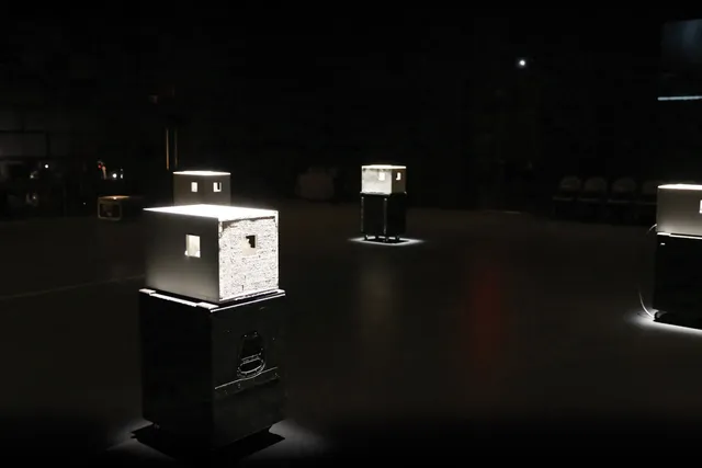 Four lit white boxes with small cutouts on black pedestals in a black box studio. 