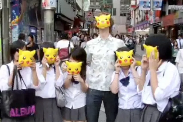 A tall white man with a group of five asian woman posing with Pikachu masks on a bustling city street. 