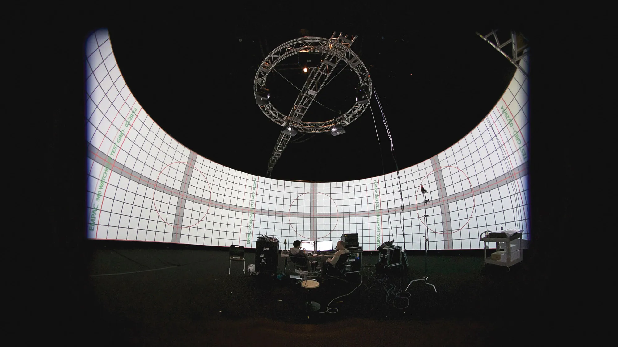 Two people sitting amongst computers and various electronics in front of a panoramic screen displaying a grid. 