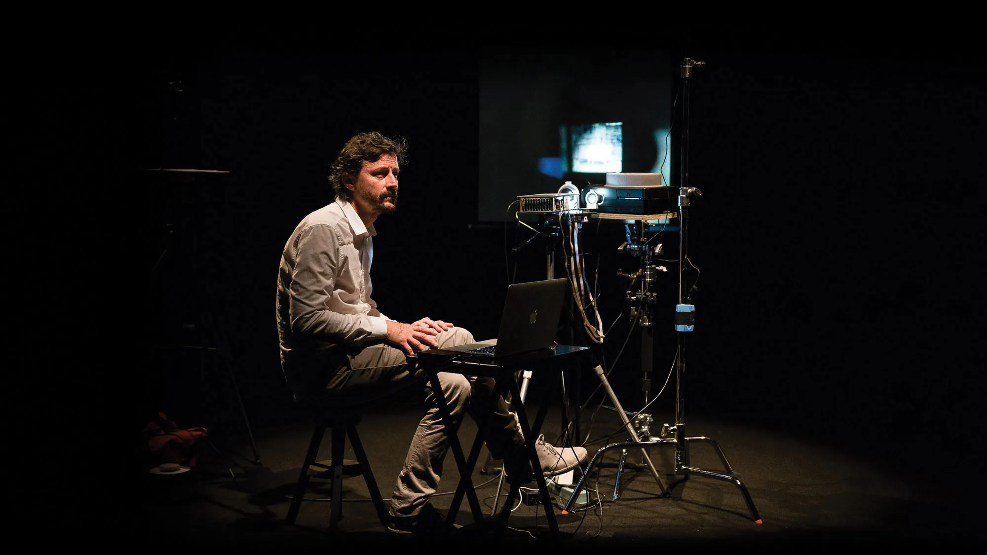 A white man with dark hair sitting cross legged next to a slide projector and lap top. 