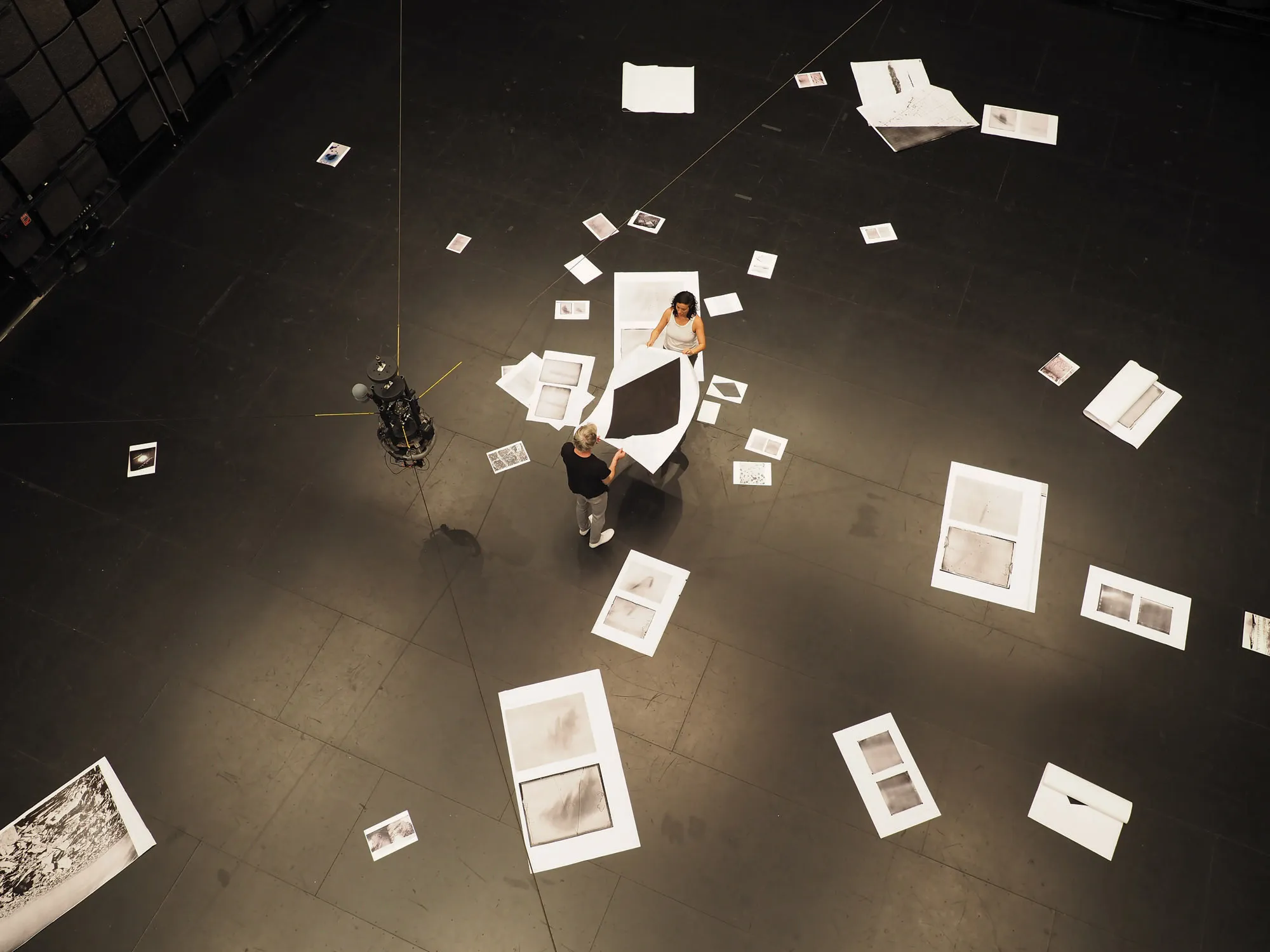 A man and a woman holding a large white piece of paper with a black diamond amongst other papers of various sizes strewn about the floor of a black box studio 