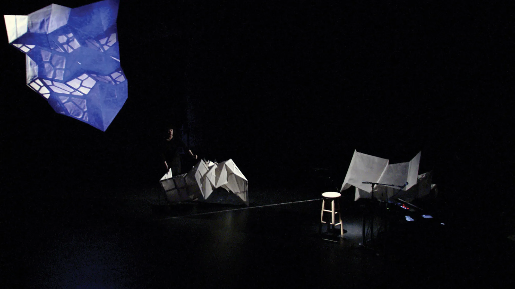 A woman on a dark stage amongst three large, white geometric sculptures. 