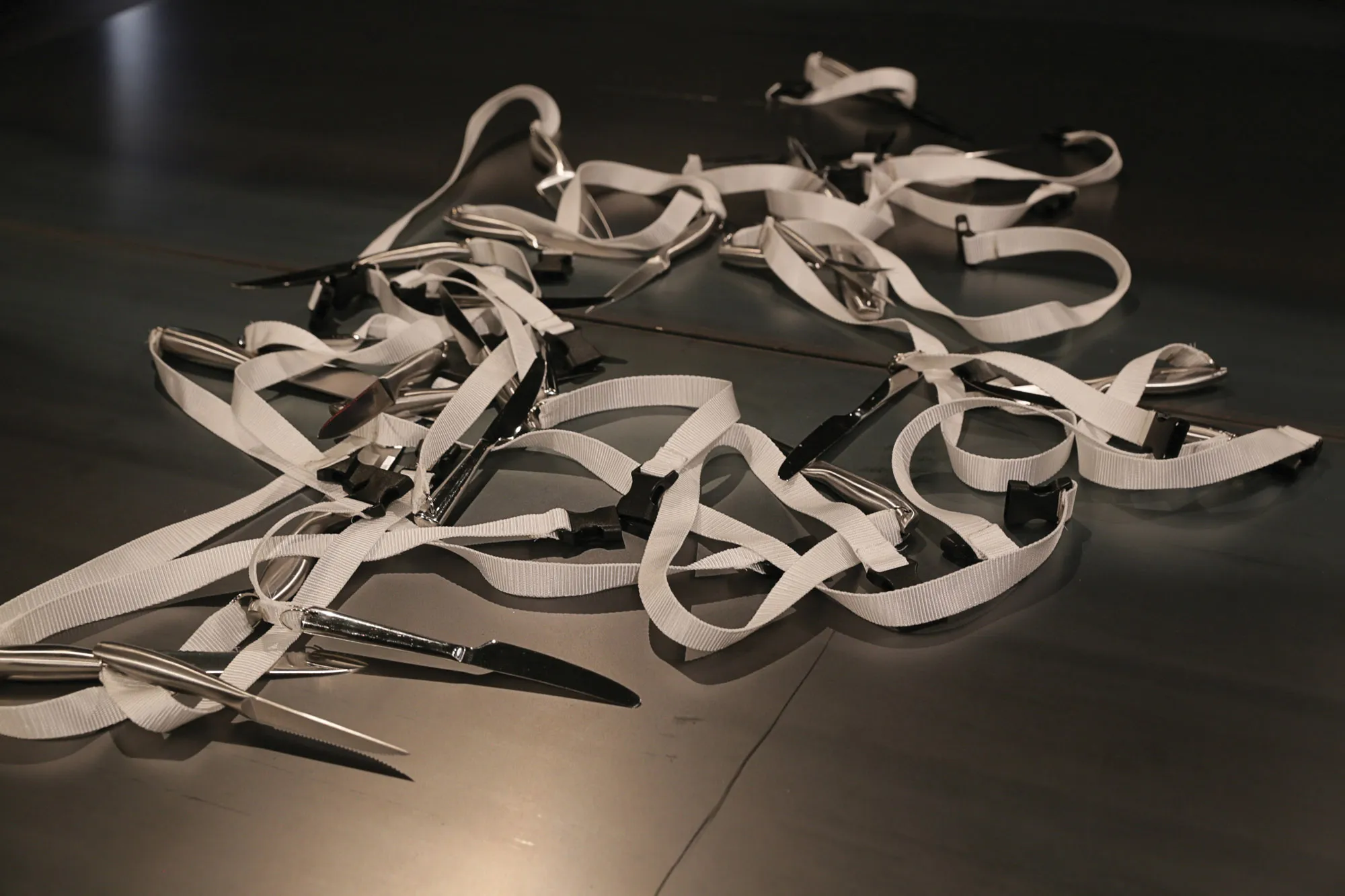 A white strap tangled with butter knives attached lying on a black floor.