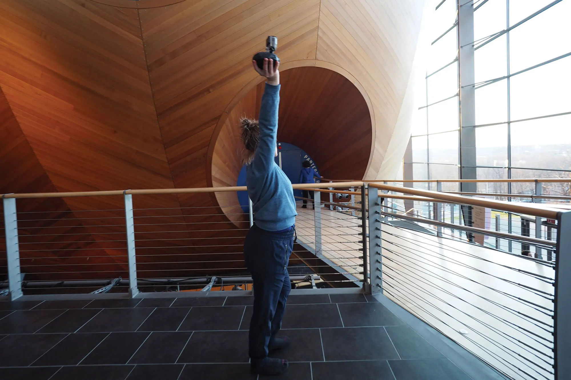 A woman in a grey sweatshirt and black joggers holding a camera above her head in front of the concert hall. 