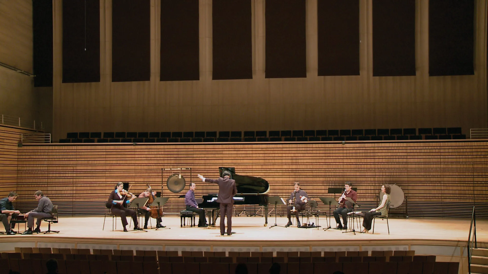 A small orchestra spread out across the concert hall stage as two men play chess in the corner of the stage. 