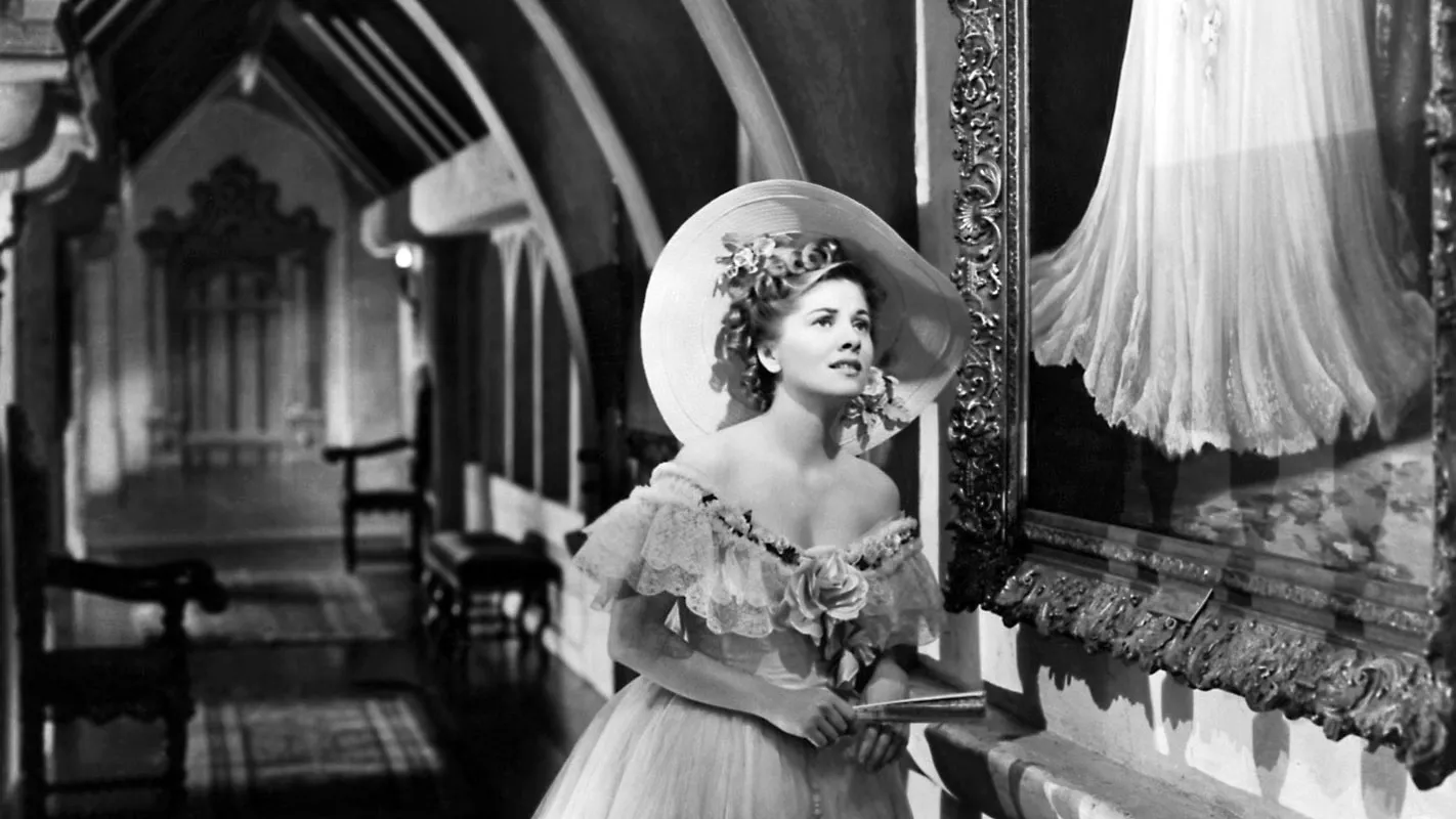 A southern woman dressed in civil war era clothing standing in an ornately decorated hallway, looking pensively at a painting. 