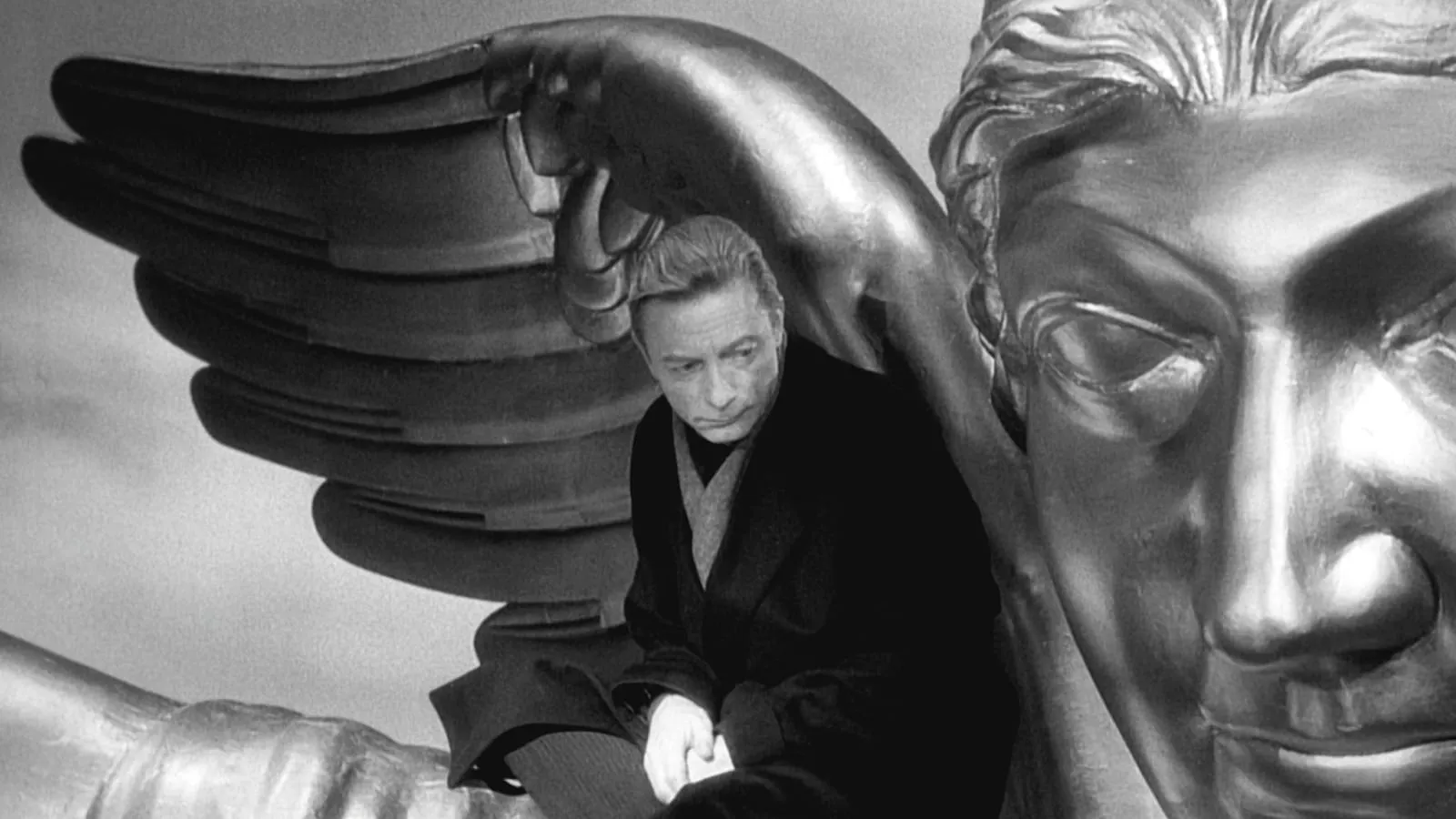 A man wearing a black overcoat looking pensively over his shoulder sitting beneath the wing of a giant metallic angel statue. 