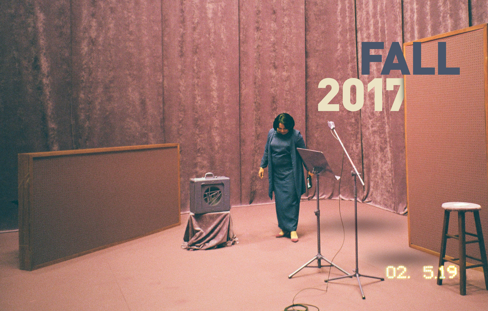 A Black woman wearing a floor length blue dress standing in all pink recording studio, complete with crushed velvet pink wall hanging and pink rug. Fall 2017 