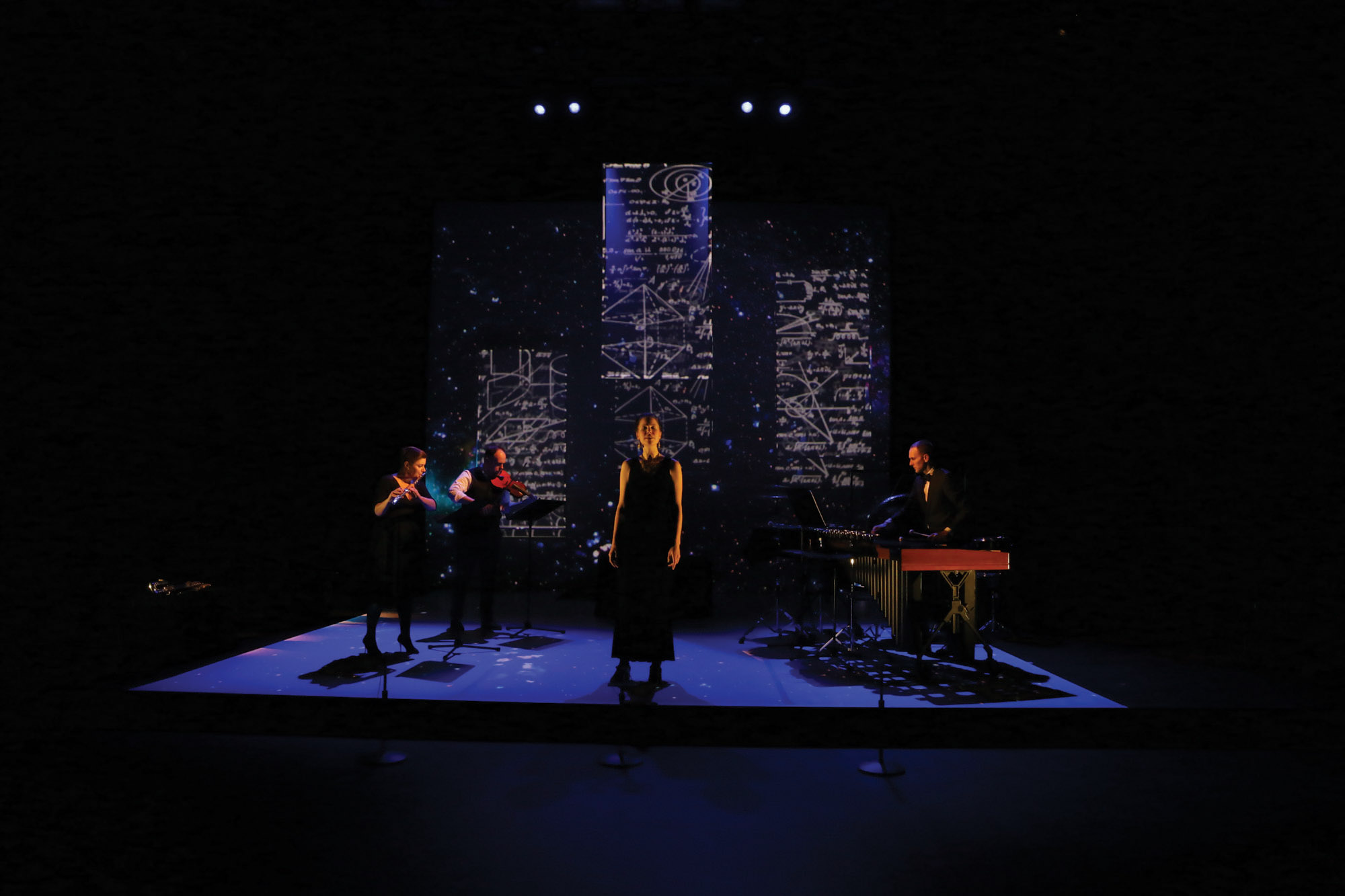 A woman standing on a dark stage lit with blue light in front of projections mathematical equations was three musicians play behind her. 
