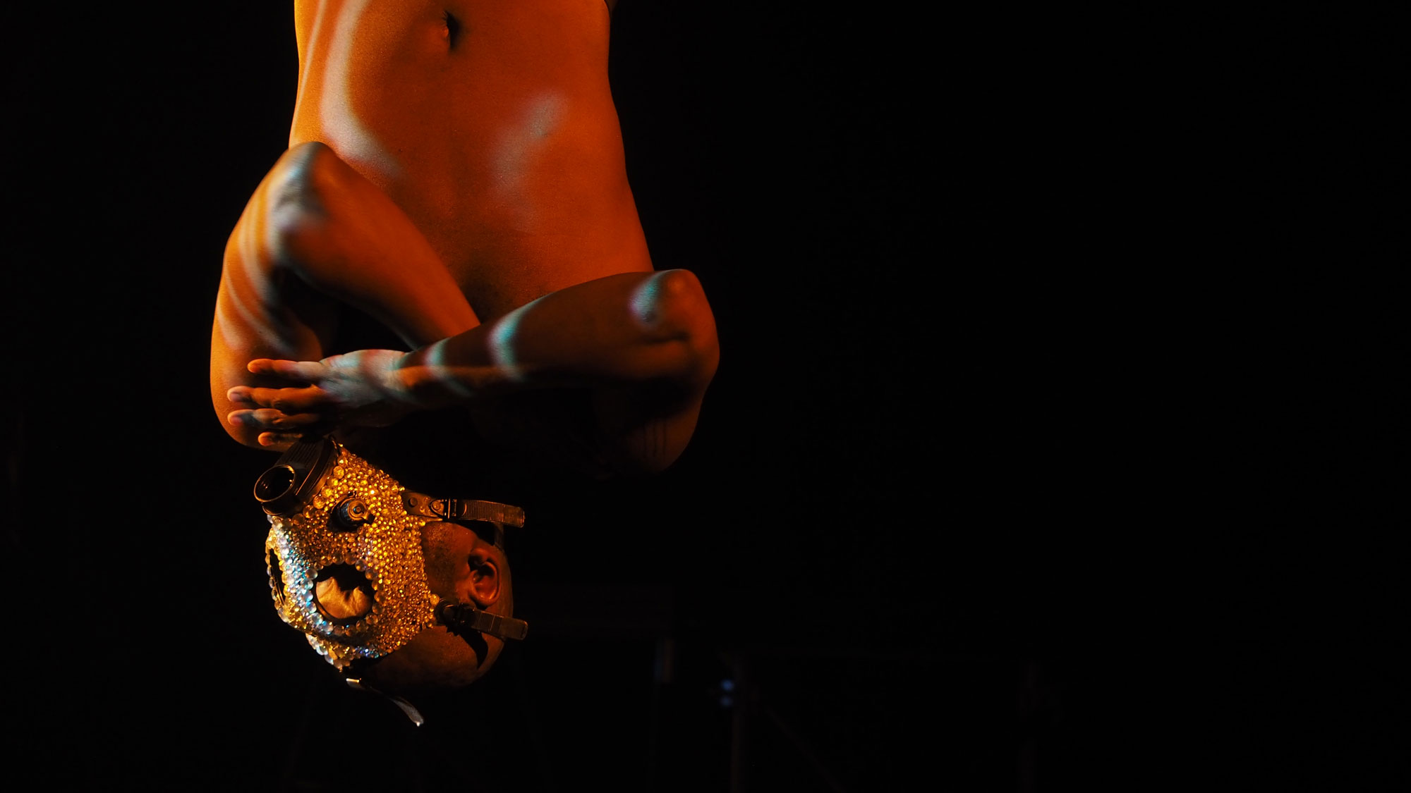 A shirtless Black man with arms crossed hanging upside down wearing a bedazzled gas mask. 