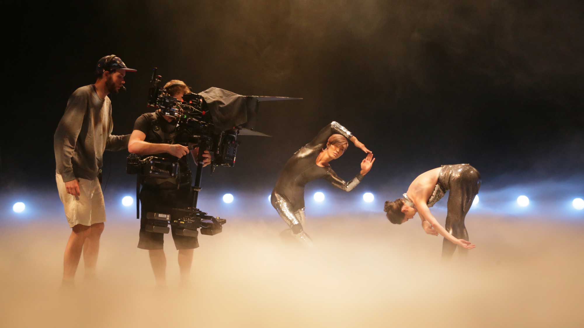 Two camera operators shooting two dancers wearing silver unitards standing in a sea of theatrical fog. 