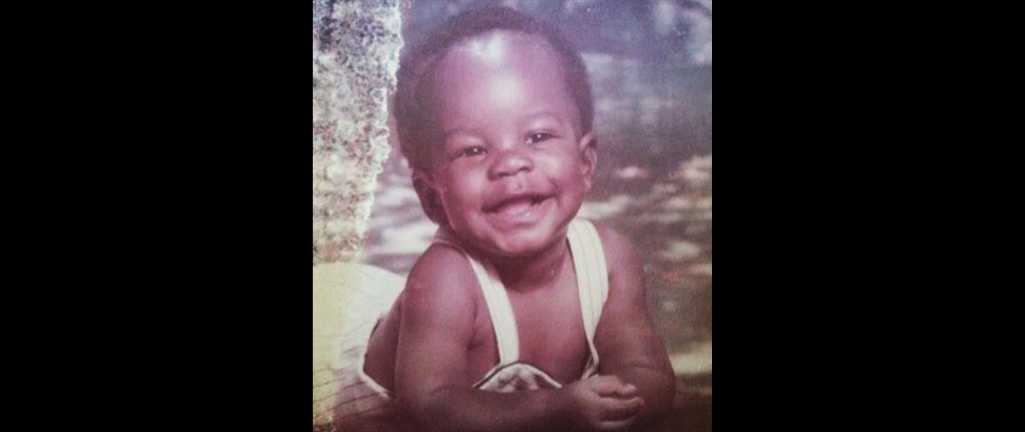 A Black baby wearing white overalls and posed in a studio portrait smiling contagiously. 