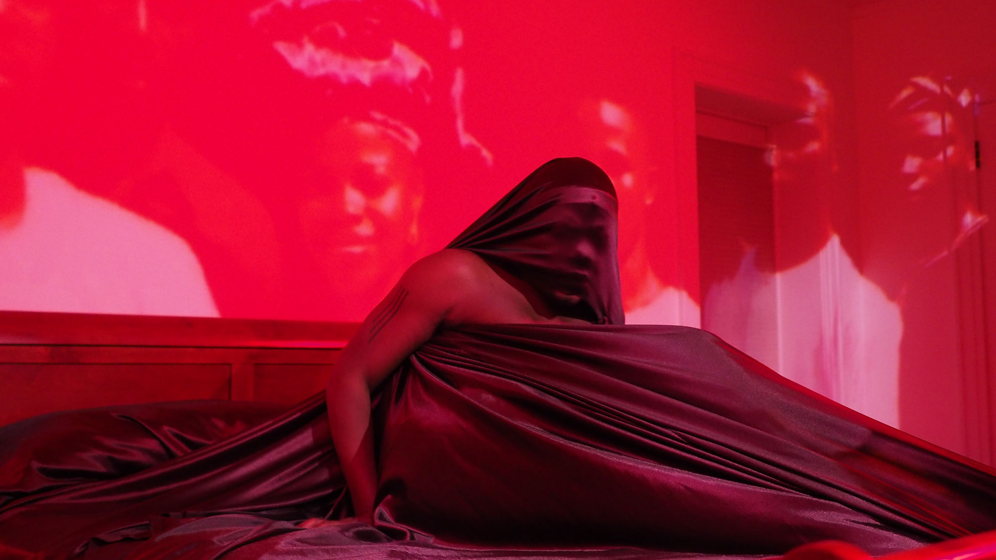 A shirtless Black man giving a guttural cry with his head covered in brown silk sheets. Images of ancestors projected behind him in a room washed in red light. 