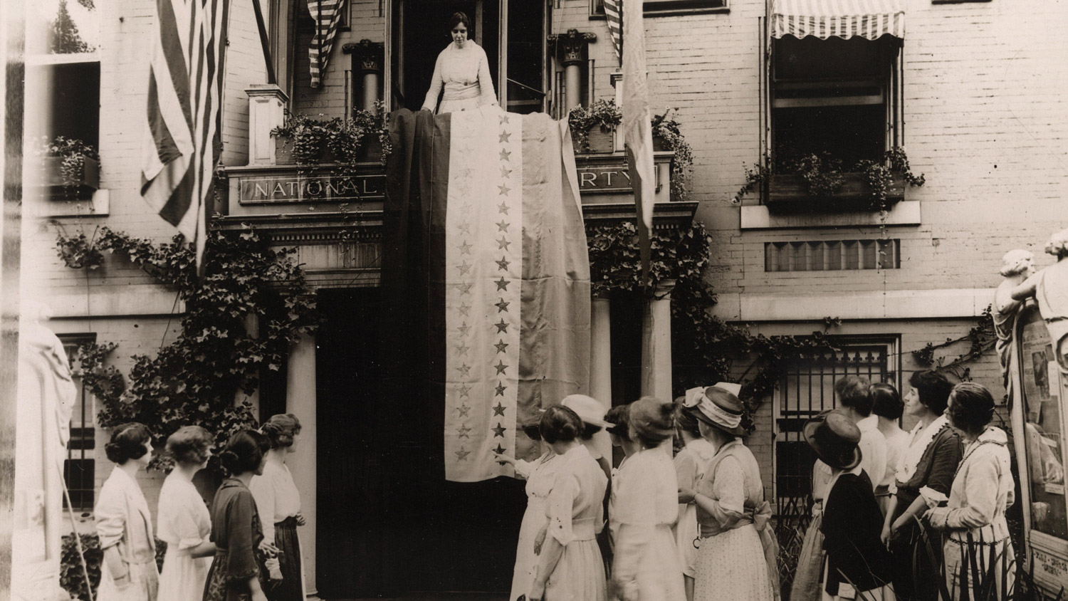 Alice Paul unfurls ratification banner off a balcony on August 18, 1920 so a crowd of period dressed woman below. 