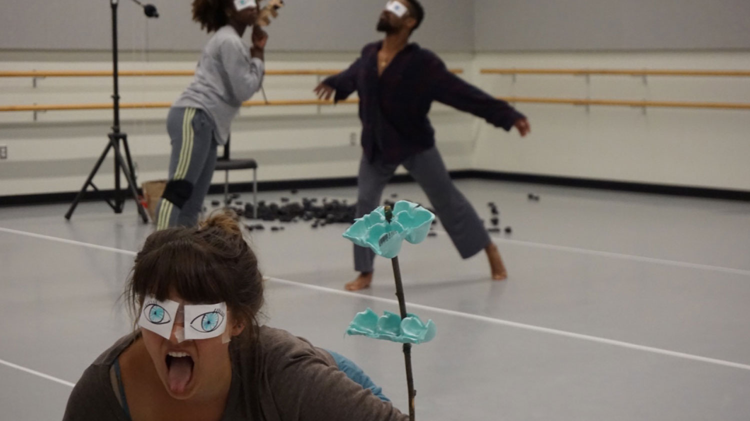 A woman with paper eyes taped over her eyes sticking out her tongue as dancers move through an empty dance studio in the background. 