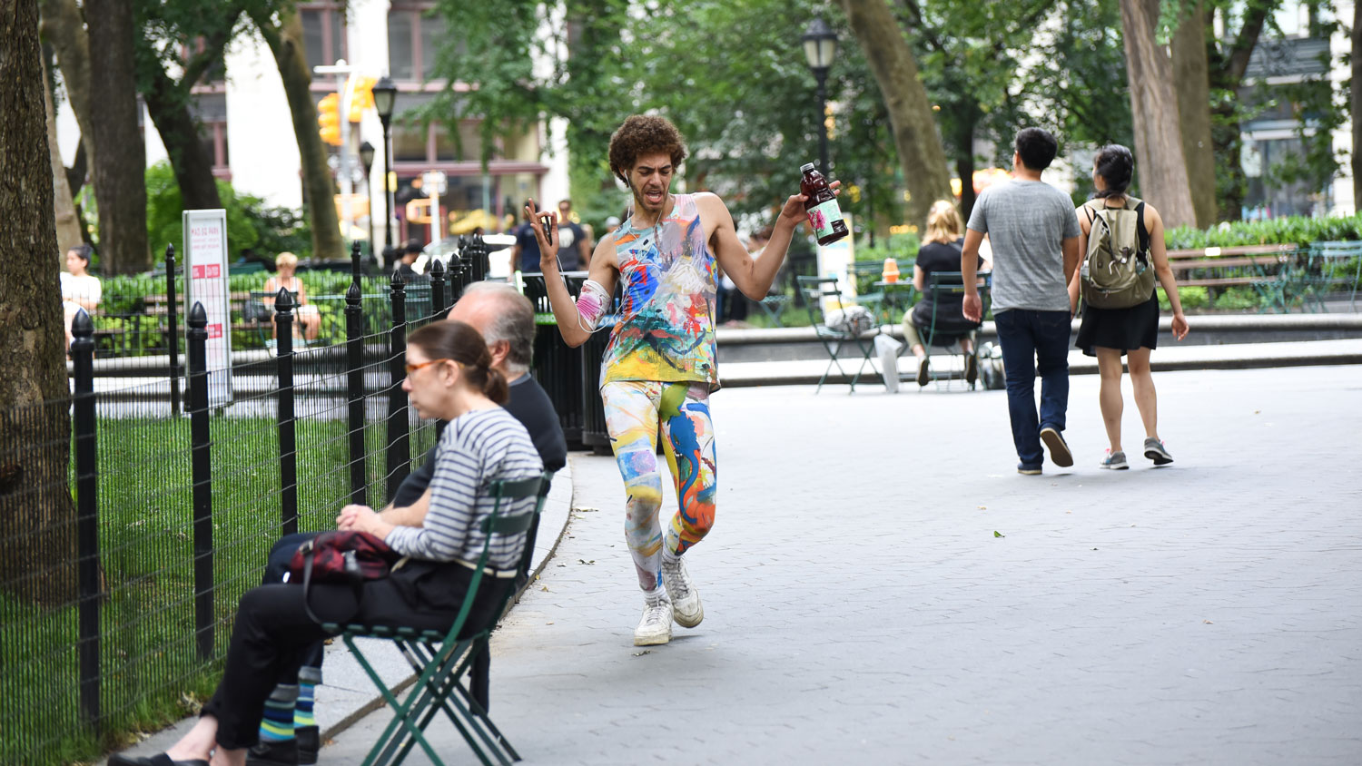 A Black man wearing rainbow paint splatted tank top and leggings strutting with headphones on in a busy New York City park. 