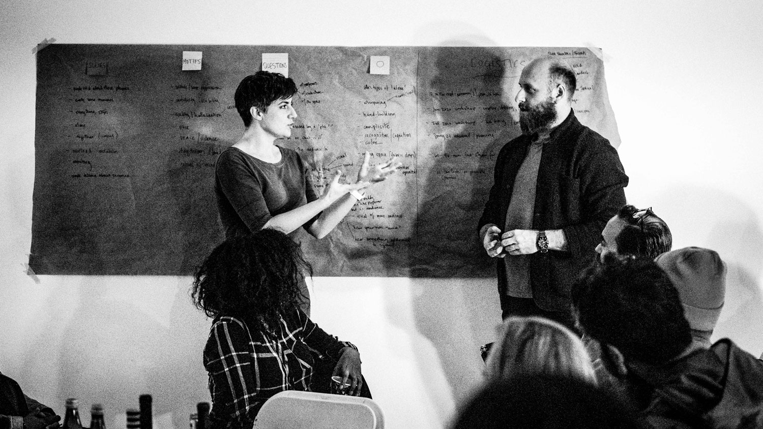 Annie Saunders talking to a bearded man in front of a blackboard in front of a small crowd. 
