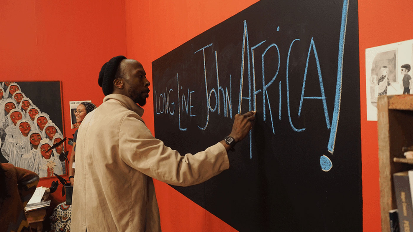 A Black man wearing a tan jacket and rolled black beanie writes on a blackboard "long live john africa!" in a red room. 
