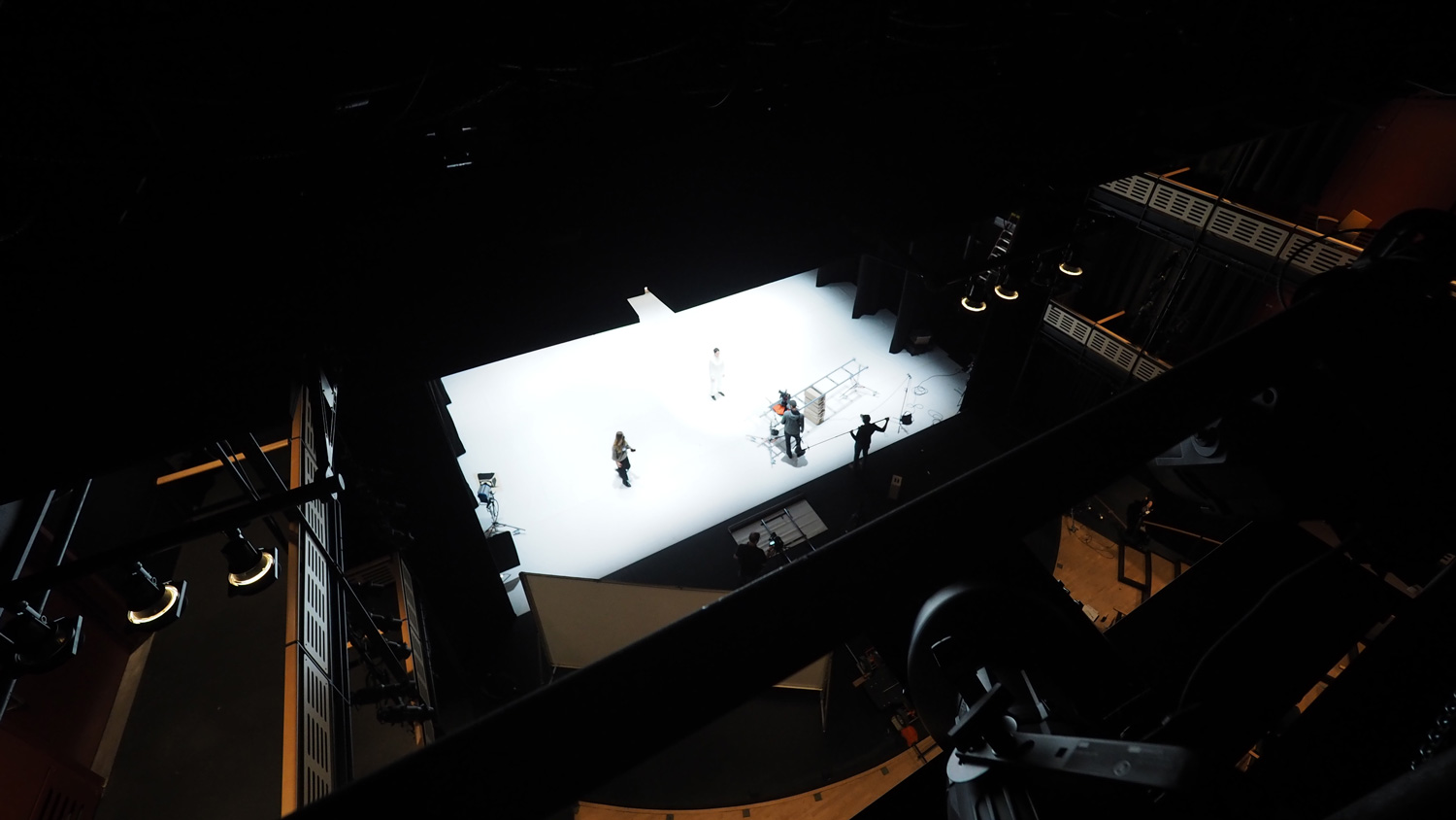 An aerial view of a dark theater with a white stage as a crew works behind the scenes. 