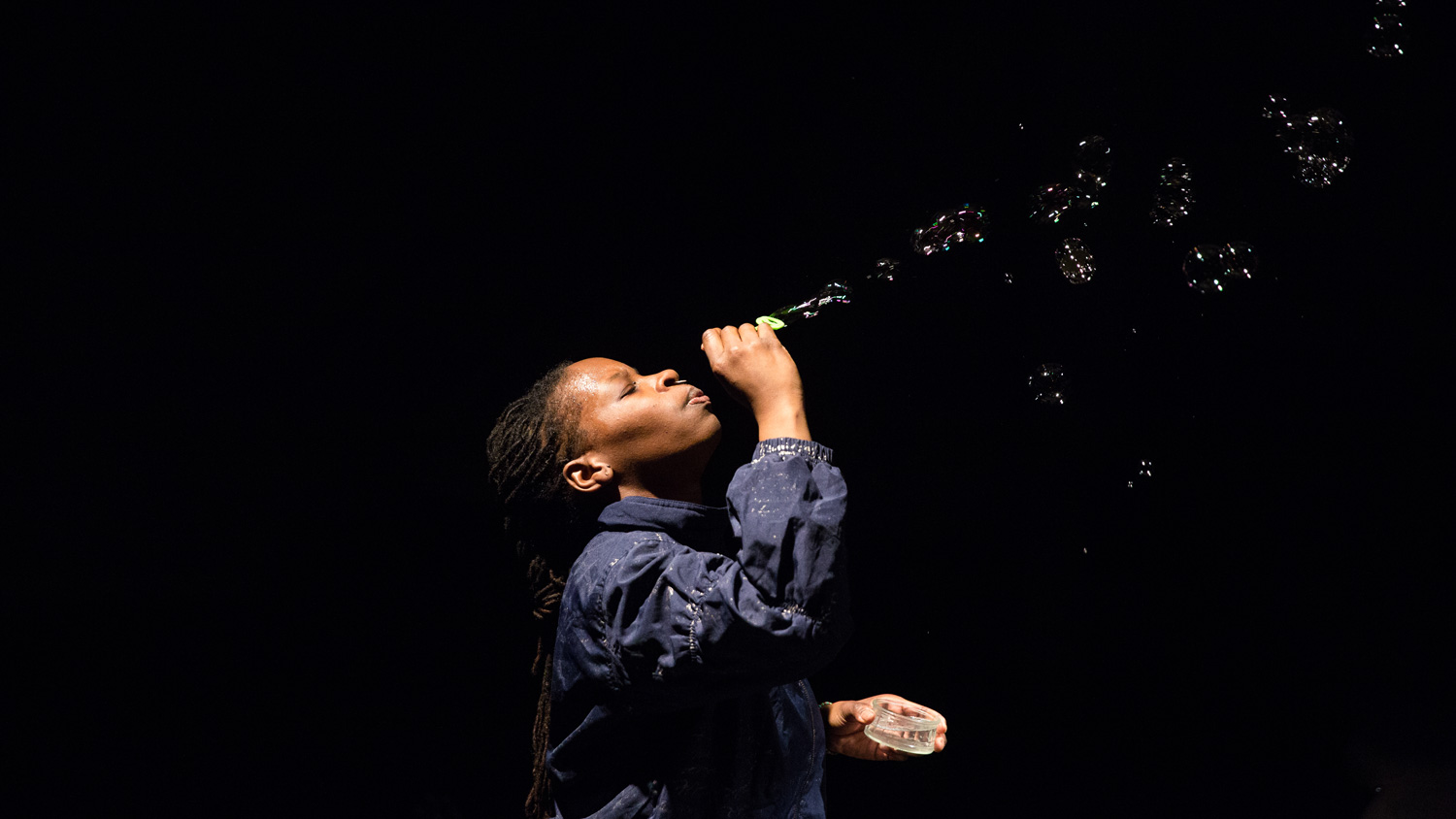 A Black person with long braided hair and wearing a blue shirt blows bubbles up into a dark room. 