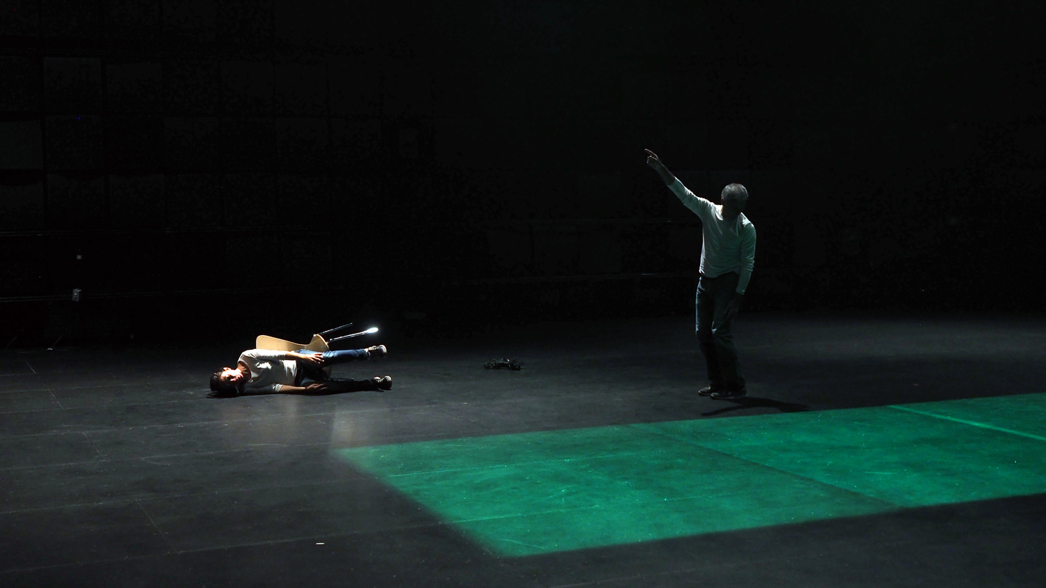 A man standing on a black stage pointing up as another person lays on the floor tipped over in their chair. A green rectangle is projected on to the floor. 