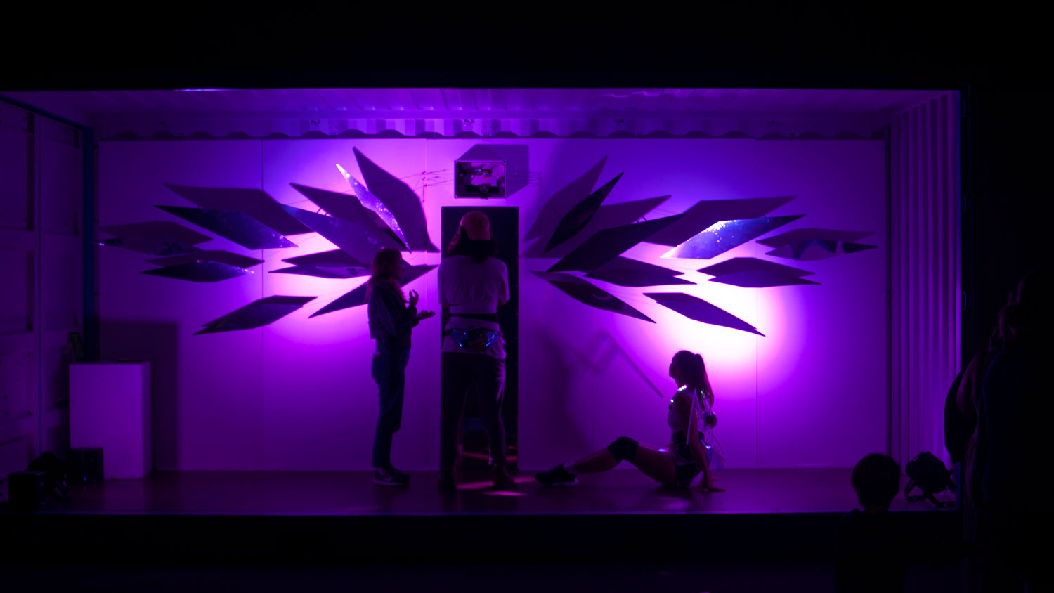 Two people on stage silohetted against a purple lit background with geometric leafy like shapes. Another person stance with back to the view in the middle of the scene, but hidden by a shadow. 