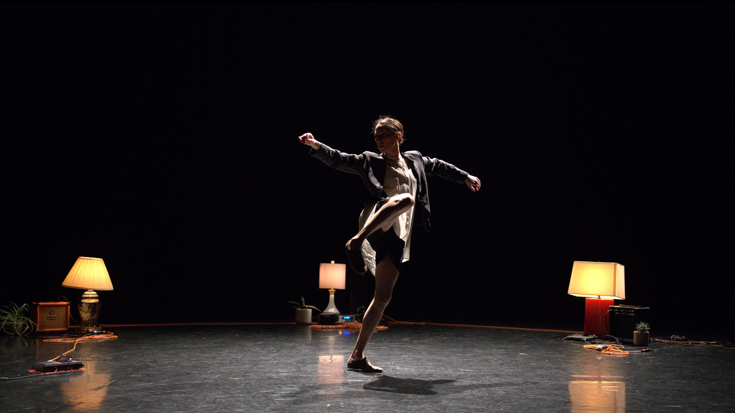 A female dancer wearing a white dress and blazer dancing with arms outstretched in a ring of orange extension cords and warmly lit lamps. 