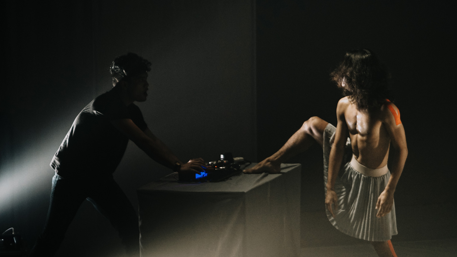 A shirtless person wearing a pleated skirt lifts their leg onto a keyboard behind controlled by a silhouetted figure on a black stage. 